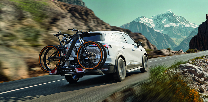Mitsubishi Grandis Zubehör – mit dem Fahrradheckträger entspannt in die Berge Weißer Mitsubishi Grandis mit montiertem Fahrradheckträger und zwei Fahrrädern fährt dynamisch auf einer Bergstraße, im Hintergrund schneebedeckte Gipfel.