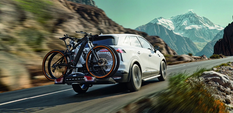 Mitsubishi Grandis Zubehör – mit dem Fahrradheckträger entspannt in die Berge Weißer Mitsubishi Grandis mit montiertem Fahrradheckträger und zwei Fahrrädern fährt dynamisch auf einer Bergstraße, im Hintergrund schneebedeckte Gipfel.