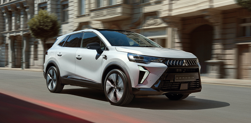 Wei&szlig;er Mitsubishi Grandis Kompakt-SUV f&auml;hrt dynamisch eine sonnige Stadtstra&szlig;e entlang, im Hintergrund Geb&auml;ude mit eleganter Fassade.