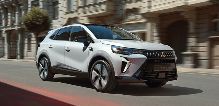 Wei&szlig;er Mitsubishi Grandis Kompakt-SUV f&auml;hrt dynamisch eine sonnige Stadtstra&szlig;e entlang, im Hintergrund Geb&auml;ude mit eleganter Fassade.