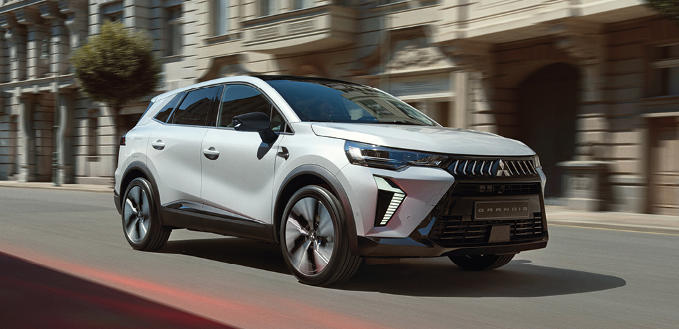 Wei&szlig;er Mitsubishi Grandis Kompakt-SUV f&auml;hrt dynamisch eine sonnige Stadtstra&szlig;e entlang, im Hintergrund Geb&auml;ude mit eleganter Fassade.