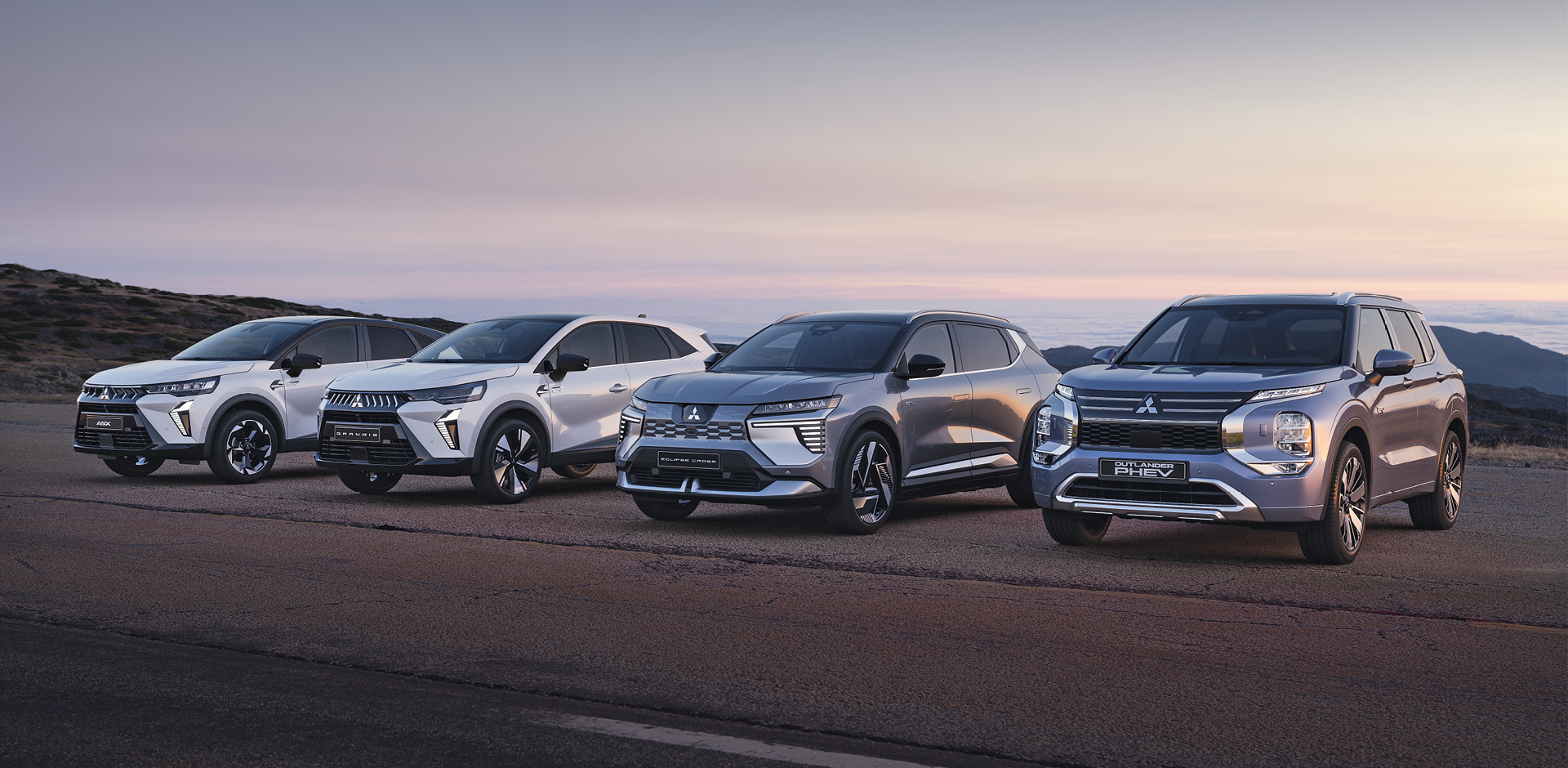 Vier Mitsubishi Modelle, darunter der Grandis, ASX, Eclipse Cross und Outlander, stehen nebeneinander auf einem offenen Platz vor weiter Landschaft bei Sonnenuntergang.