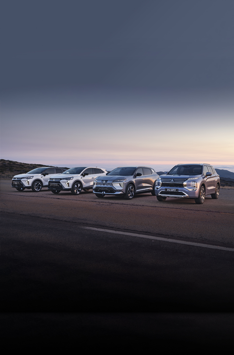 Vier Mitsubishi Modelle, darunter der Grandis, ASX, Eclipse Cross und Outlander, stehen nebeneinander auf einem offenen Platz vor weiter Landschaft bei Sonnenuntergang.