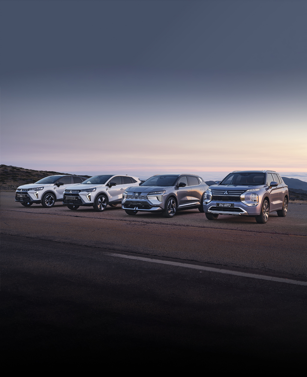 Vier Mitsubishi Modelle, darunter der Grandis, ASX, Eclipse Cross und Outlander, stehen nebeneinander auf einem offenen Platz vor weiter Landschaft bei Sonnenuntergang.