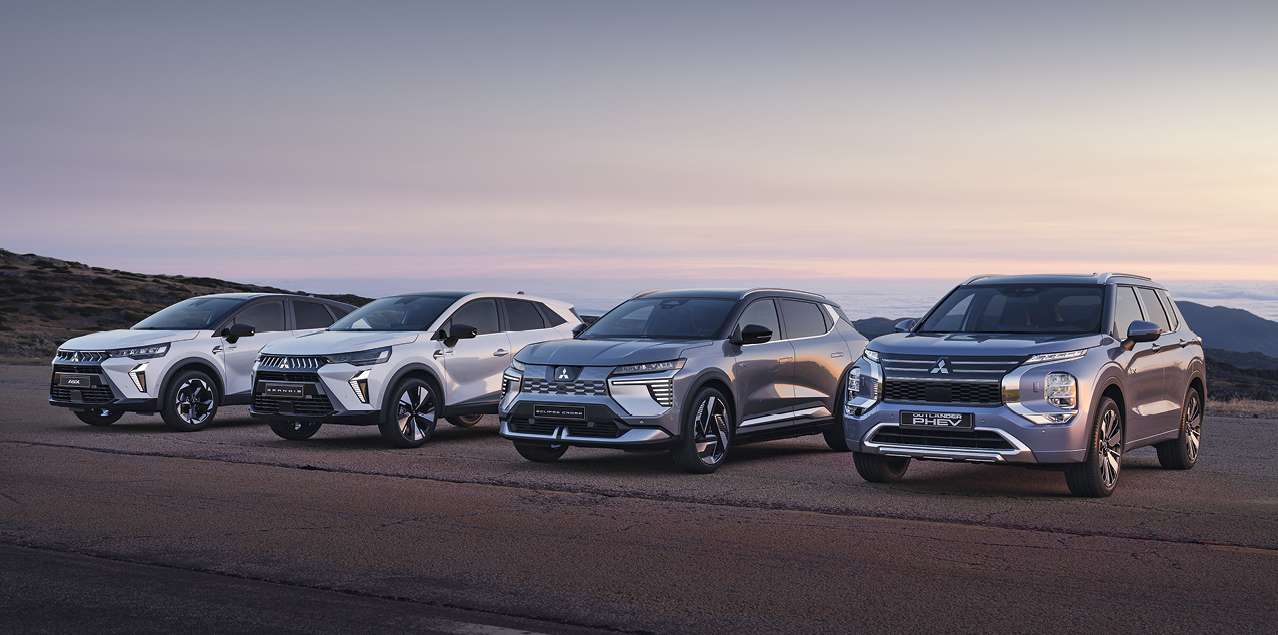 Vier Mitsubishi Modelle, darunter der Grandis, ASX, Eclipse Cross und Outlander, stehen nebeneinander auf einem offenen Platz vor weiter Landschaft bei Sonnenuntergang.