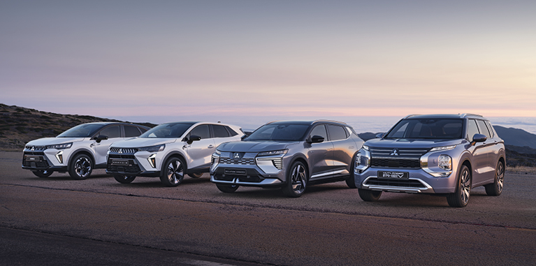 Vier Mitsubishi Modelle, darunter der Grandis, ASX, Eclipse Cross und Outlander, stehen nebeneinander auf einem offenen Platz vor weiter Landschaft bei Sonnenuntergang.
