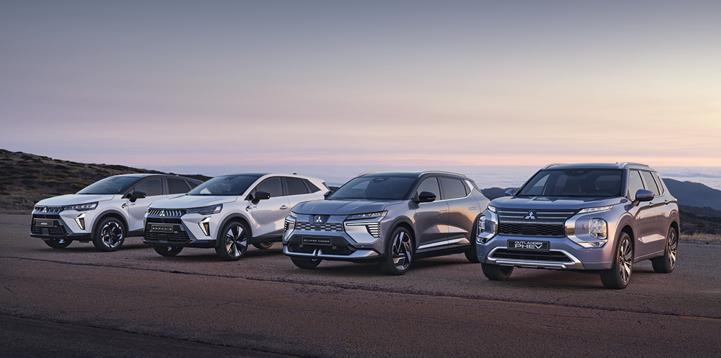 Vier Mitsubishi Modelle, darunter der Grandis, ASX, Eclipse Cross und Outlander, stehen nebeneinander auf einem offenen Platz vor weiter Landschaft bei Sonnenuntergang.