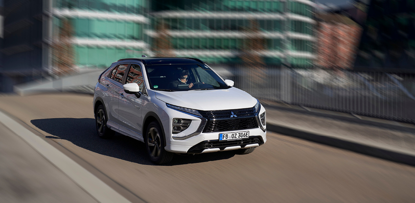 Wei&szlig;er Mitsubishi Eclipse Cross Plug-in Hybrid f&auml;hrt auf einer Stra&szlig;e durch ein modernes Stadtviertel.