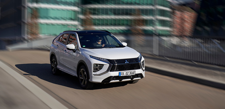 Wei&szlig;er Mitsubishi Eclipse Cross Plug-in Hybrid f&auml;hrt auf einer Stra&szlig;e durch ein modernes Stadtviertel.