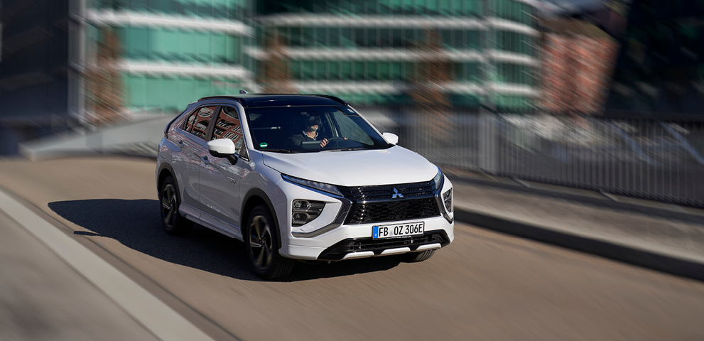 Wei&szlig;er Mitsubishi Eclipse Cross Plug-in Hybrid f&auml;hrt auf einer Stra&szlig;e durch ein modernes Stadtviertel.