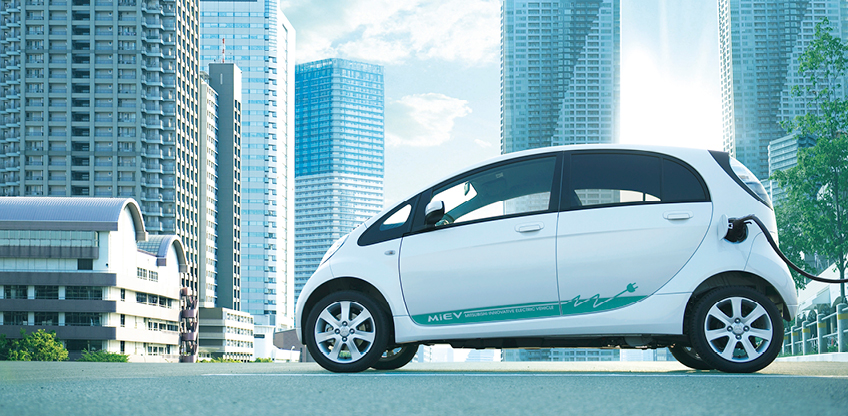 Wei&szlig;er Mitsubishi i-MiEV ist an eine Ladestation angeschlossen und steht vor einer modernen Stadtkulisse.