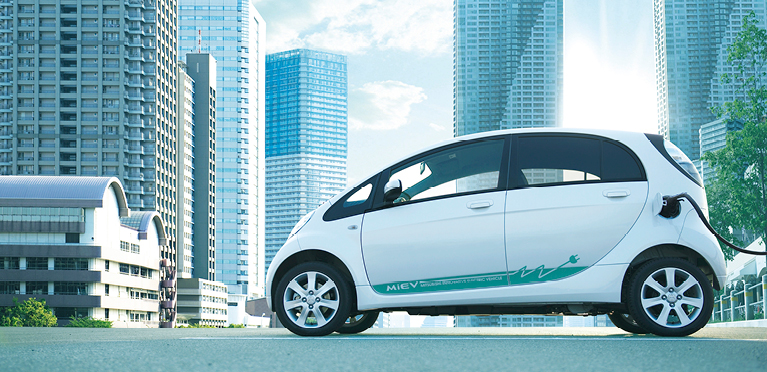 Wei&szlig;er Mitsubishi i-MiEV ist an eine Ladestation angeschlossen und steht vor einer modernen Stadtkulisse.