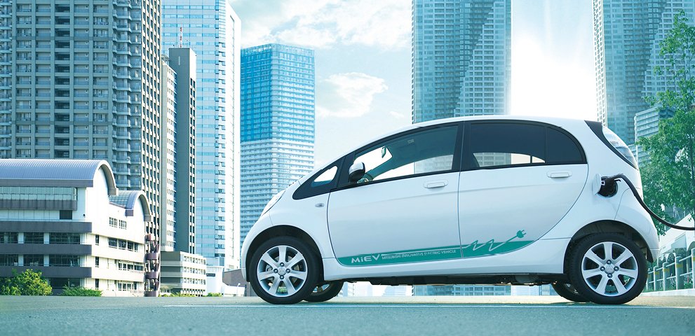 Wei&szlig;er Mitsubishi i-MiEV ist an eine Ladestation angeschlossen und steht vor einer modernen Stadtkulisse.
