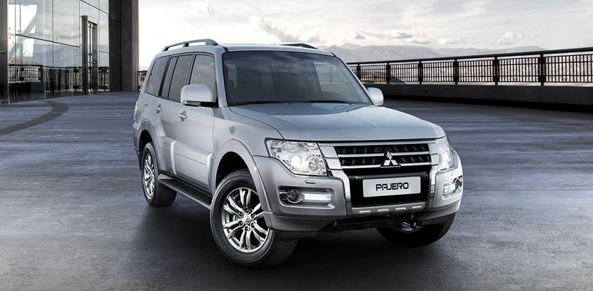 Silberner Mitsubishi Pajero steht auf einem offenen Vorplatz vor einem modernen Geb&auml;ude; im Hintergrund sind Berge zu sehen.