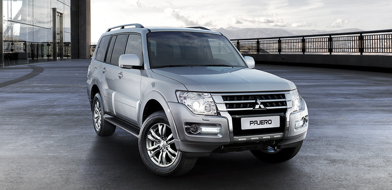 Silberner Mitsubishi Pajero steht auf einem offenen Vorplatz vor einem modernen Geb&auml;ude; im Hintergrund sind Berge zu sehen.