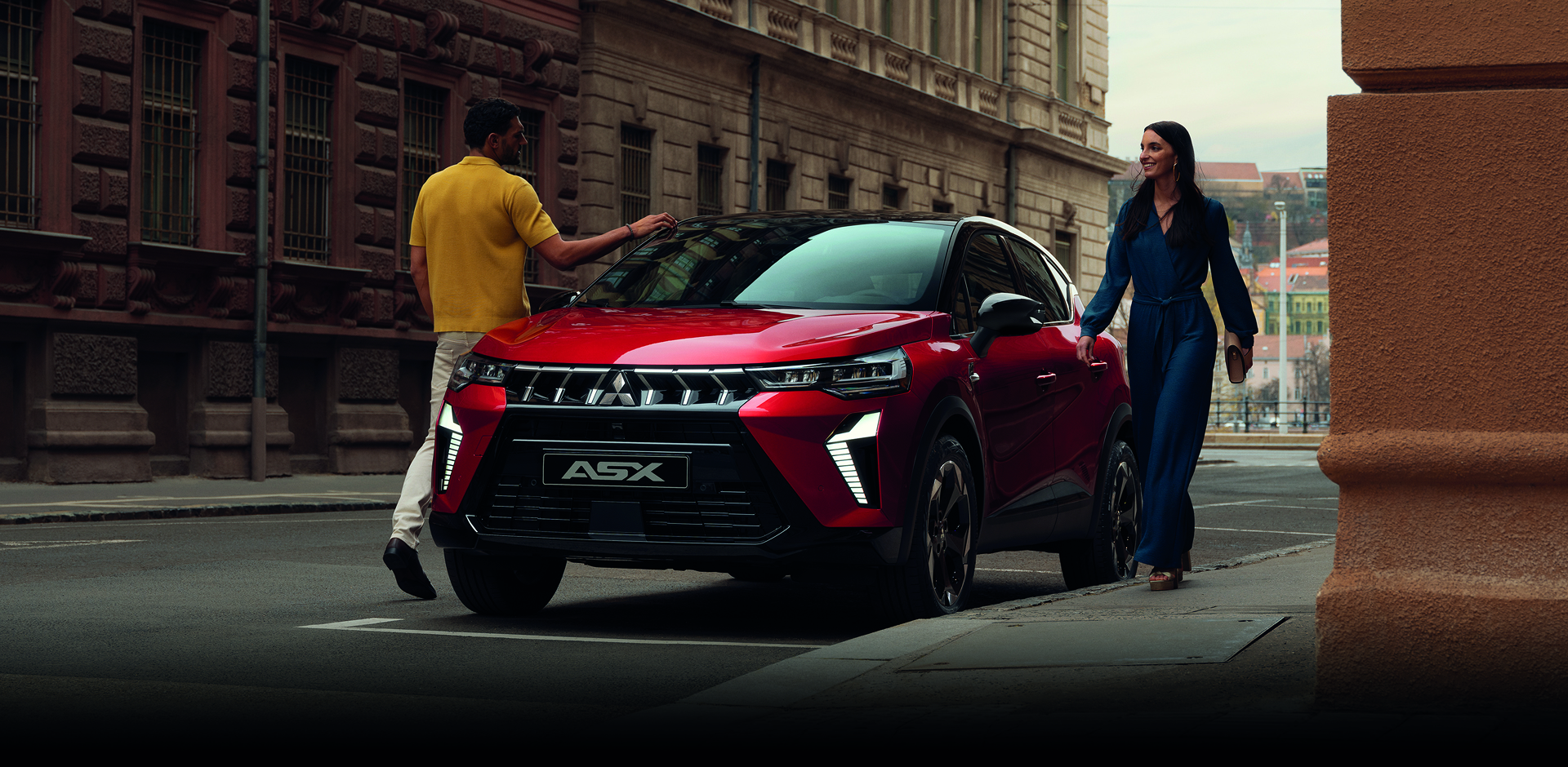 Roter Mitsubishi ASX steht in einer Stadtstra&szlig;e, ein Mann und eine Frau gehen l&auml;chelnd am Fahrzeug vorbei &ndash; Fokus auf dem markanten Frontdesign.