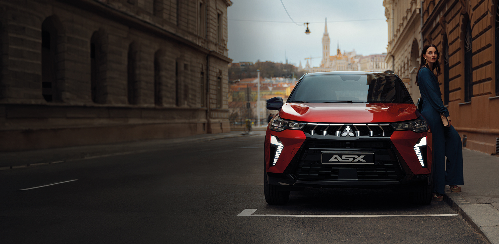 Roter Misubishi ASX SUV steht frontal auf einer Stadtstra&szlig;e, moderne LED-Lichtsignatur und K&uuml;hlergrill im Fokus, rechts daneben eine Frau, die sich an das Fahrzeug lehnt.