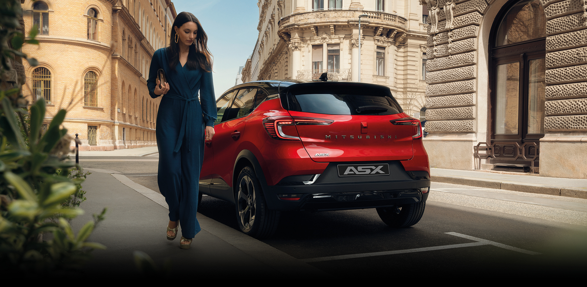 Roter Mitsubishi ASX Kompakt-SUV von hinten auf einer eleganten Stadtstra&szlig;e, moderne R&uuml;ckleuchten und Heckdesign im Fokus, daneben geht eine Frau im blauen Jumpsuit.