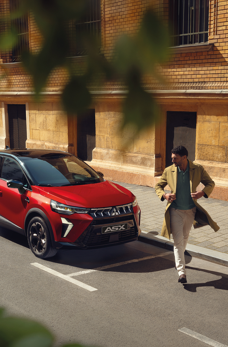 Roter Mitsubishi ASX Kompakt-SUV parkt in der Stadt, Front- und Seitenansicht; vor dem Fahrzeug &uuml;berquert ein Mann in beigem Mantel die Stra&szlig;e.