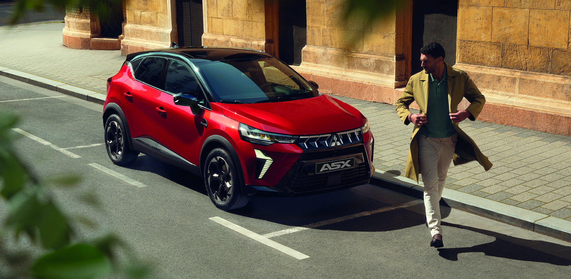 Roter Mitsubishi ASX SUV steht am Stra&szlig;enrand in der Stadt, ein Mann in beigem Mantel geht vor dem Fahrzeug &uuml;ber die Stra&szlig;e.