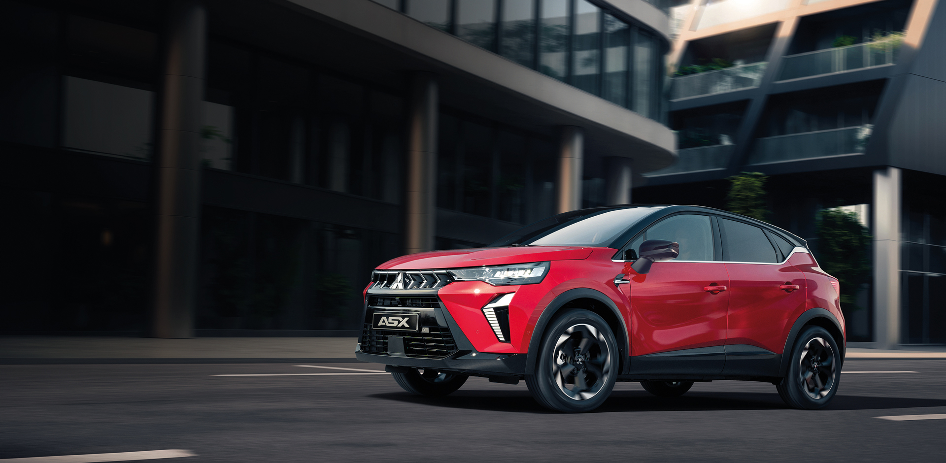 Roter Mitsubishi ASX Kompakt-SUV f&auml;hrt dynamisch durch die Stadt vor einem modernen Geb&auml;ude.