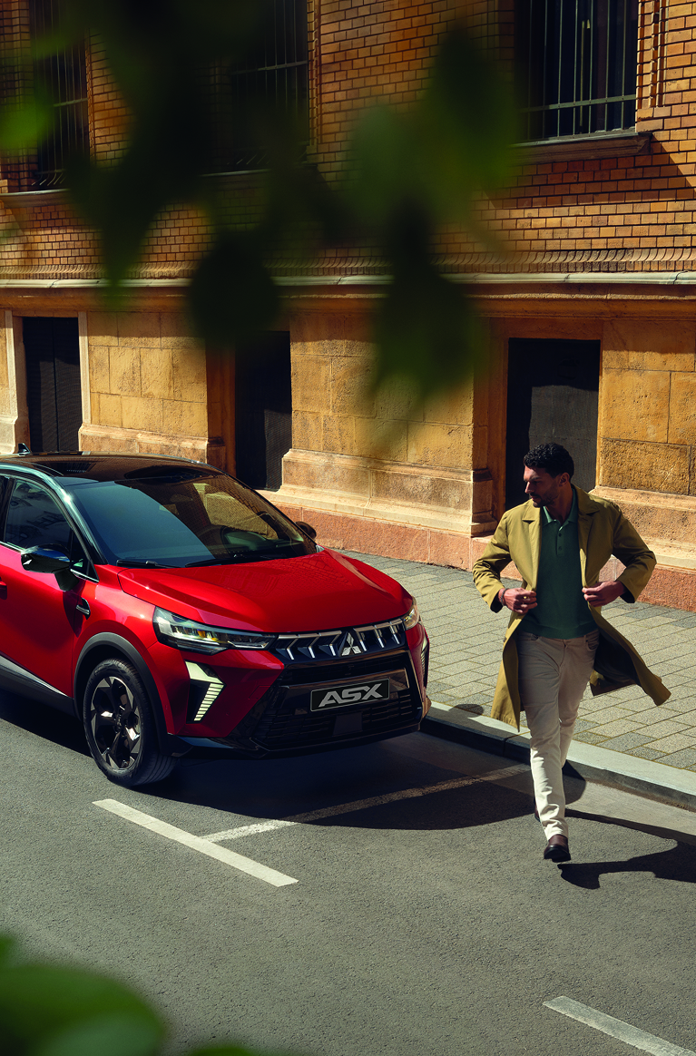 Roter Mitsubishi ASX parkt an einer sonnigen Stadtstra&szlig;e vor einem historischen Geb&auml;ude; ein elegant gekleideter Mann geht entspannt am Fahrzeug vorbei.