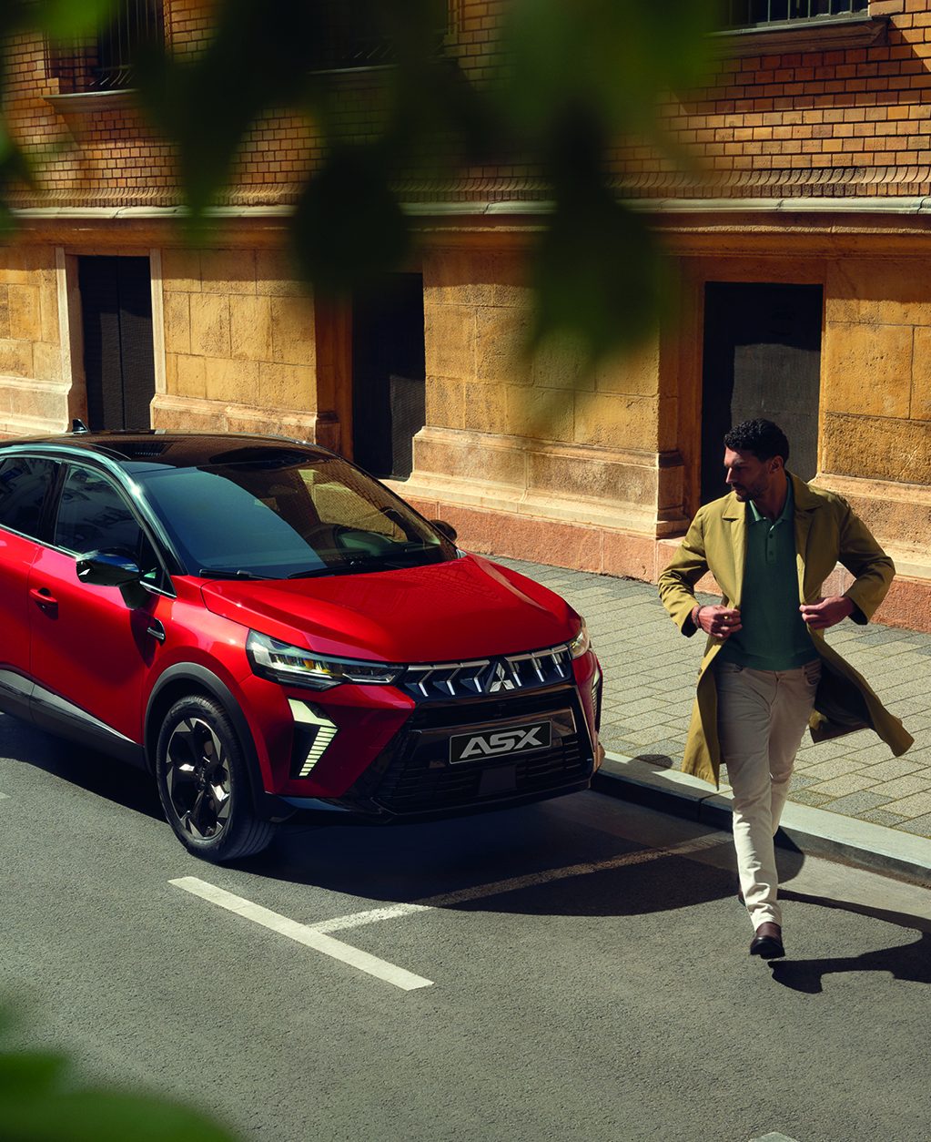 Roter Mitsubishi ASX parkt an einer sonnigen Stadtstra&szlig;e vor einem historischen Geb&auml;ude; ein elegant gekleideter Mann geht entspannt am Fahrzeug vorbei.