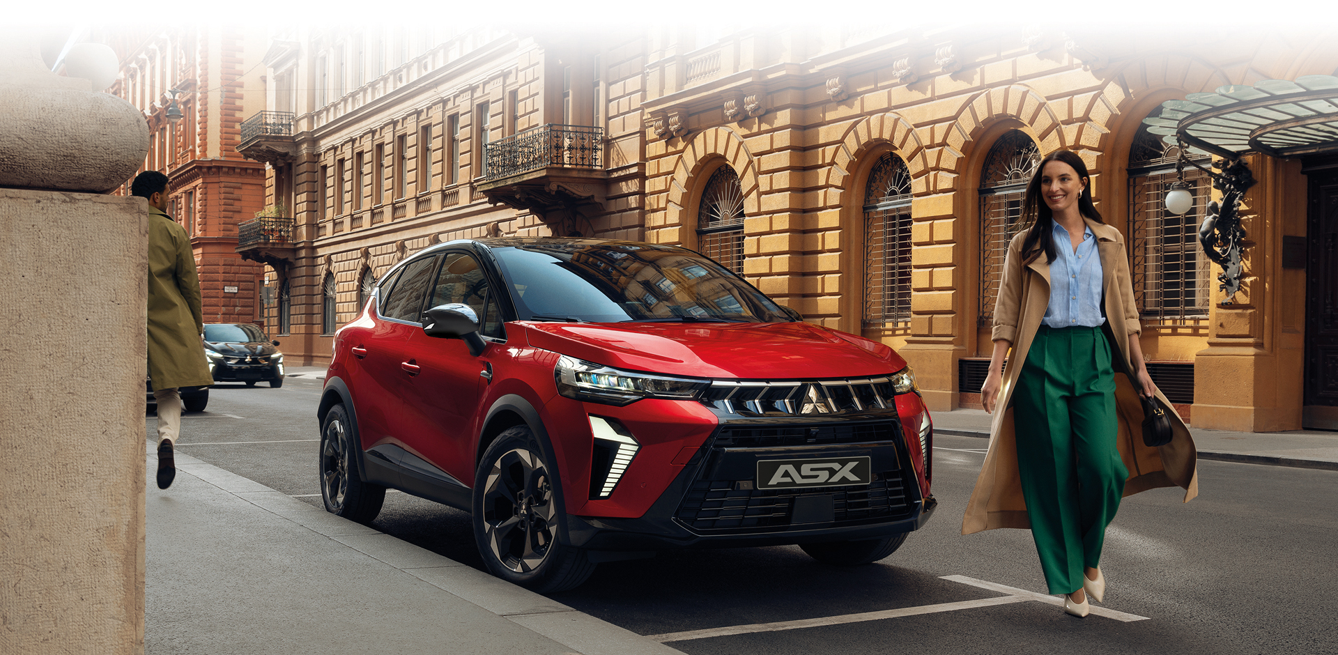 Roter Mitsubishi ASX in Front Seitenansicht auf einer Stadtstra&szlig;e vor historischen Geb&auml;uden, eine Frau geht l&auml;chelnd am Fahrzeug vorbei.