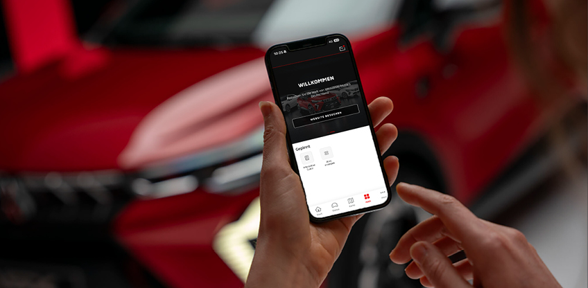 Nahaufnahme einer Hand, die ein Smartphone mit ge&ouml;ffneter MITSUBISHI MOTORS App h&auml;lt; auf dem Display ist ein Willkommen-Screen zu sehen; im Hintergrund unscharf der Mitsubishi ASX.