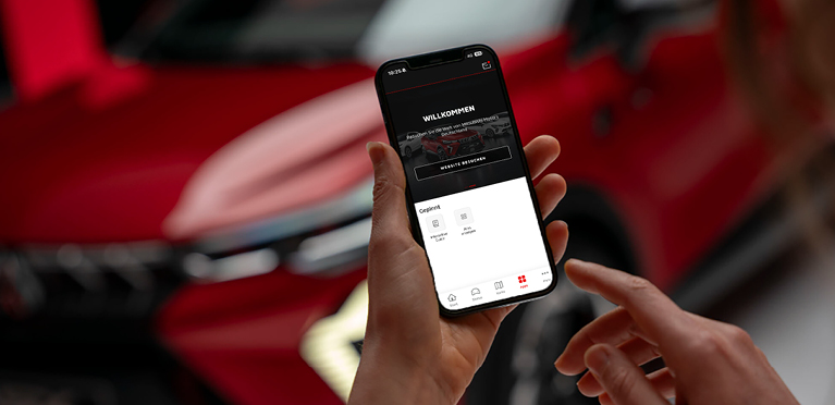 Nahaufnahme einer Hand, die ein Smartphone mit ge&ouml;ffneter MITSUBISHI MOTORS App h&auml;lt; auf dem Display ist ein Willkommen-Screen zu sehen; im Hintergrund unscharf der Mitsubishi ASX.