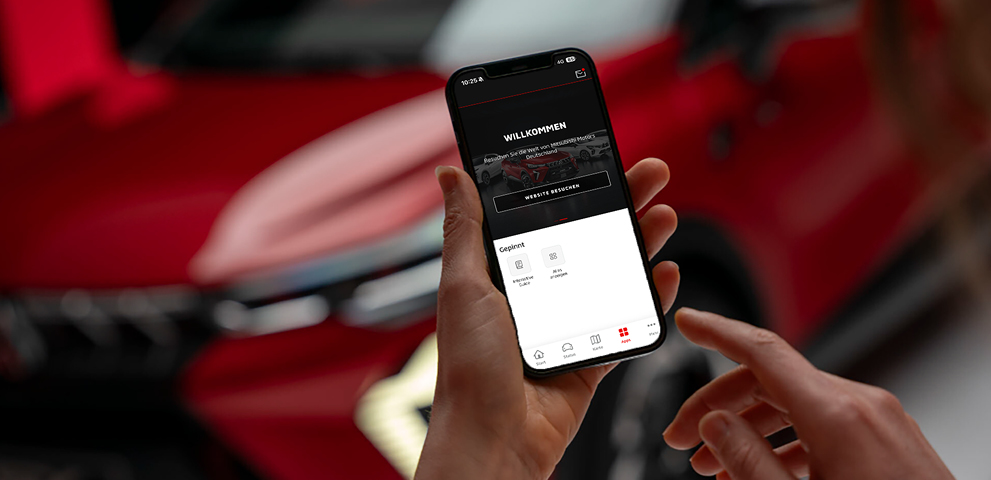 Nahaufnahme einer Hand, die ein Smartphone mit ge&ouml;ffneter MITSUBISHI MOTORS App h&auml;lt; auf dem Display ist ein Willkommen-Screen zu sehen; im Hintergrund unscharf der Mitsubishi ASX.
