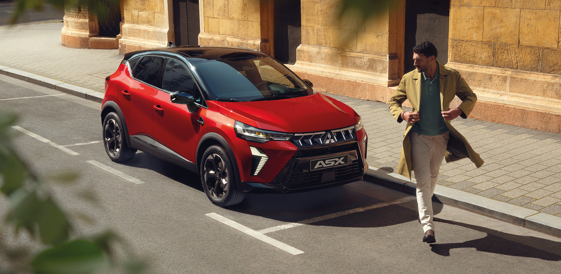 Roter Mitsubishi ASX parkt an einer sonnigen Stadtstra&szlig;e vor einem historischen Geb&auml;ude, ein Mann geht mit Mantel und Smartphone in der Hand am Fahrzeug vorbei.