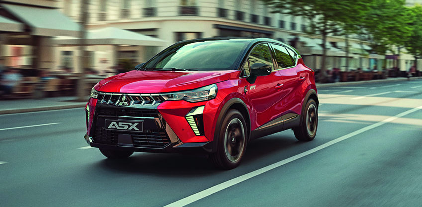 Roter Mitsubishi ASX Select in schr&auml;ger Front Seitenansicht f&auml;hrt dynamisch &uuml;ber einen breiten Stadtboulevard, im Hintergrund Stra&szlig;encaf&eacute;s, Fu&szlig;g&auml;nger und B&auml;ume &ndash; betont das moderne, urbane Design des kompakten SUV.