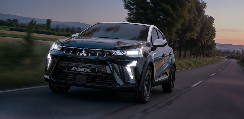 Mitsubishi ASX Black Sondermodell f&auml;hrt in der D&auml;mmerung auf einer Landstra&szlig;e, Front Seitenansicht mit eingeschalteten LED Scheinwerfern; die Szene betont den sportlich exklusiven Charakter der ASX Sondermodelle.