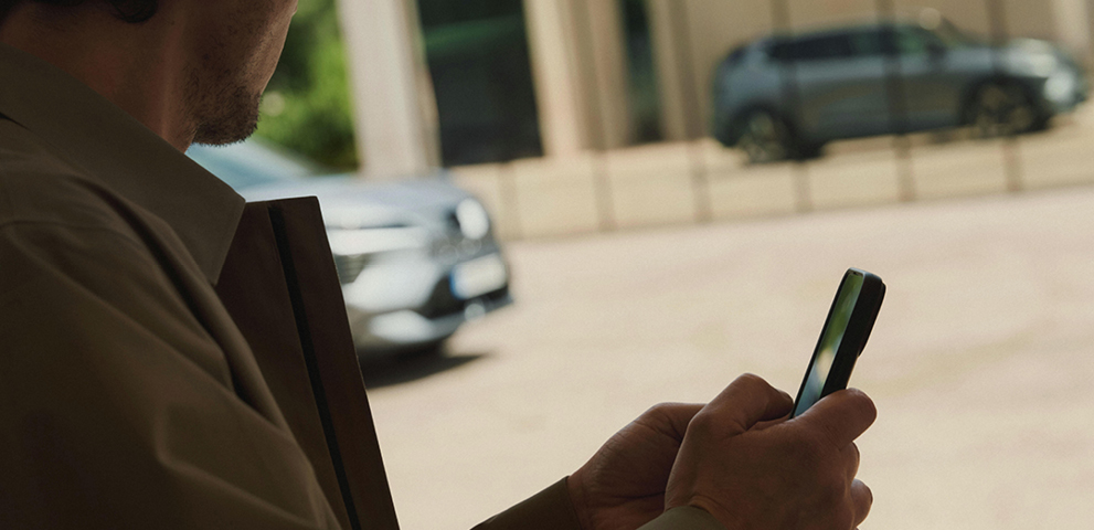 Eine Person bedient ein Smartphone, w&auml;hrend im Hintergrund ein grauer Mitsubishi Eclipse Cross zu sehen ist. Er spiegelt sich in einer Glasfront.