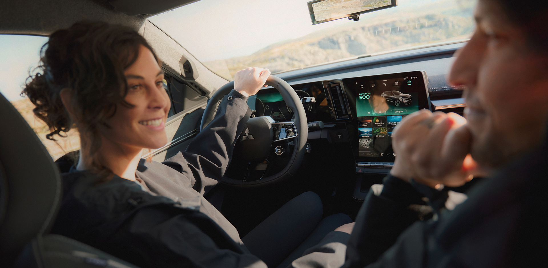 Eine Frau schaut auf dem Fahrersitz sitzend zu ihrem männlichen Beifahrer, auf dem Infotainment erkennt man die Fahreinstellung ECO.