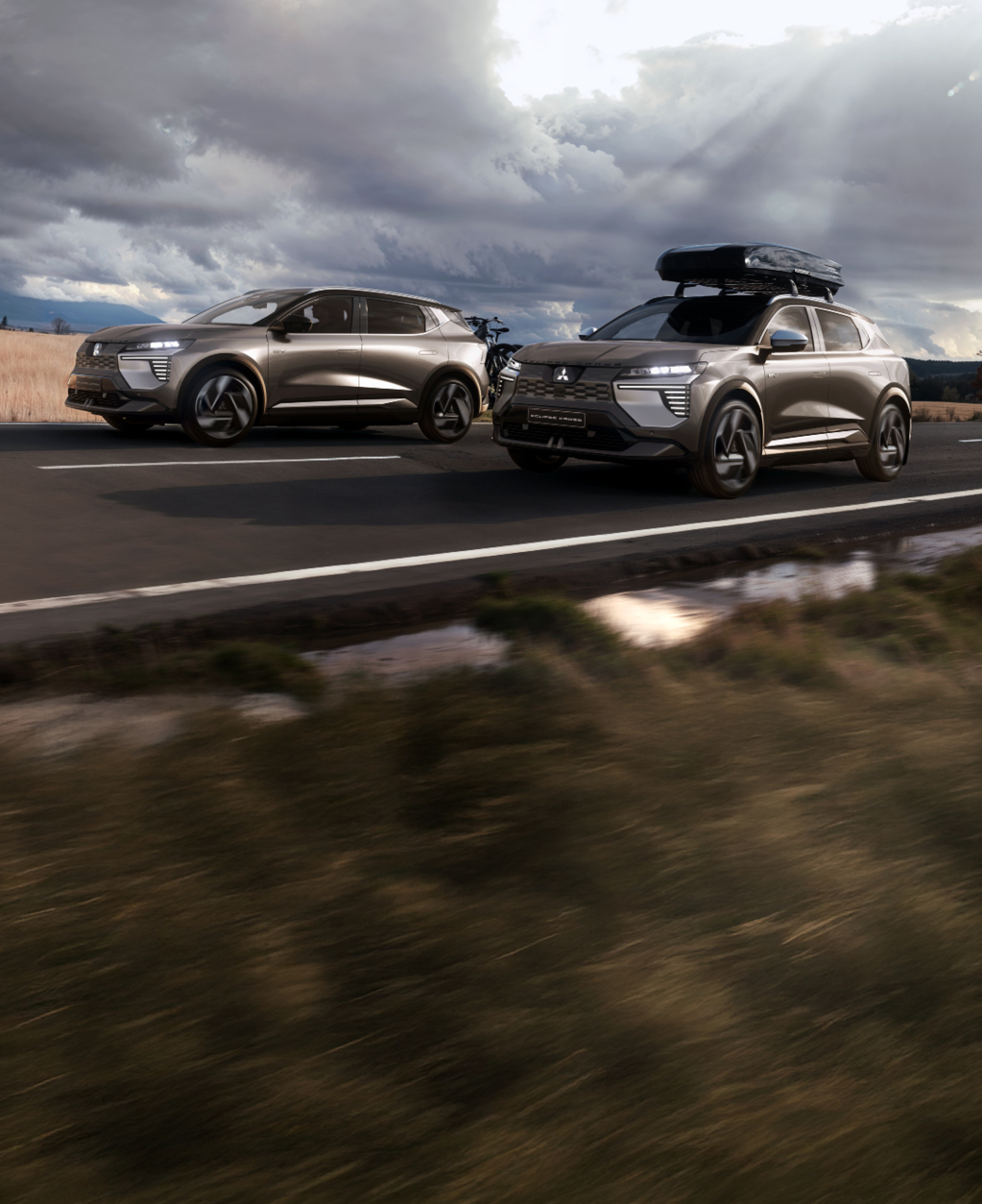 Zwei Mitsubishi Eclipse Cross, einer mit Fahrrad-Hecktr&auml;ger, einer mit Dachbox, fahren eine Landstra&szlig;e entlang.