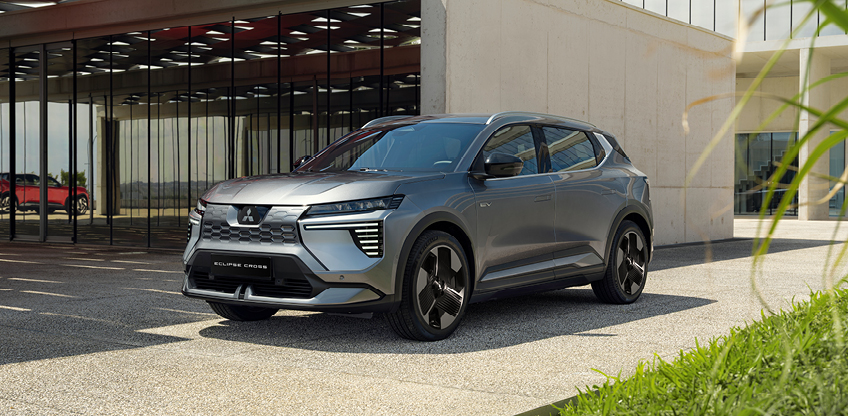 Mitsubishi Eclipse Cross Intro Edition in der Einfahrt Graue Mitsubishi Eclipse Cross Intro Edition steht in Dreiviertelansicht vor einem modernen Gebäude mit Glasfassade; im Vordergrund sind Gras und Pflanzen zu sehen.