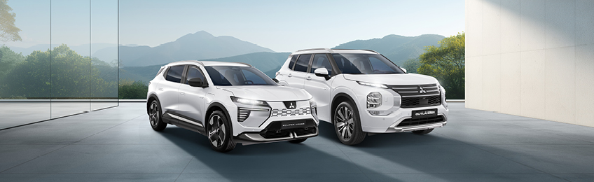 MITSUBISHI MOTORS Elektromobilität Zwei weiße Mitsubishi-SUVs - ein Outlander und ein Eclipse Cross - stehen nebeneinander in einem modernen, lichtdurchfluteten Showroom mit Blick auf eine Berglandschaft.