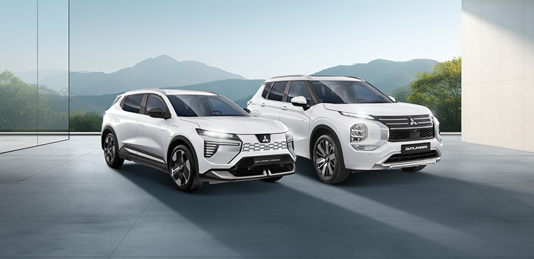 MITSUBISHI MOTORS Elektromobilität Zwei weiße Mitsubishi-SUVs - ein Outlander und ein Eclipse Cross - stehen nebeneinander in einem modernen, lichtdurchfluteten Showroom mit Blick auf eine Berglandschaft.