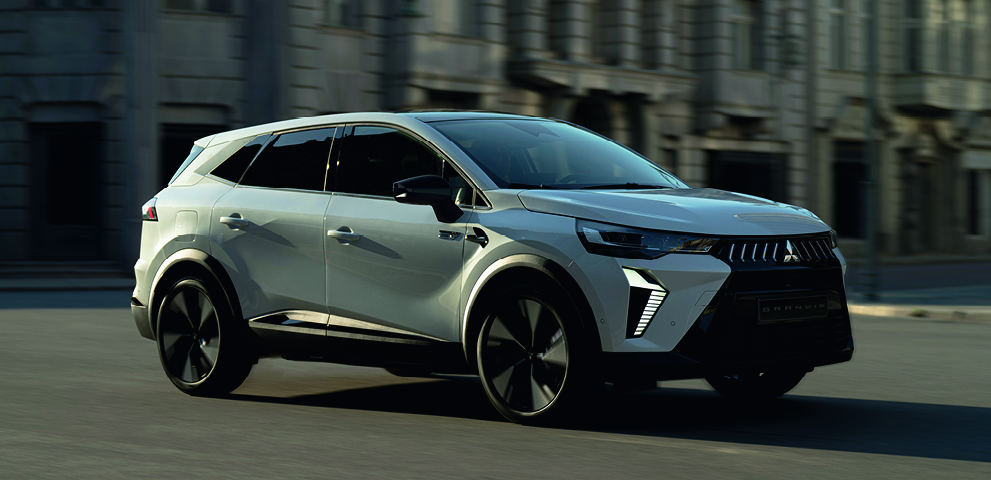 Wei&szlig;er Mitsubishi Grandis SUV f&auml;hrt dynamisch durch eine Stadt, dreiviertel Frontansicht mit unscharfem Hintergrund.