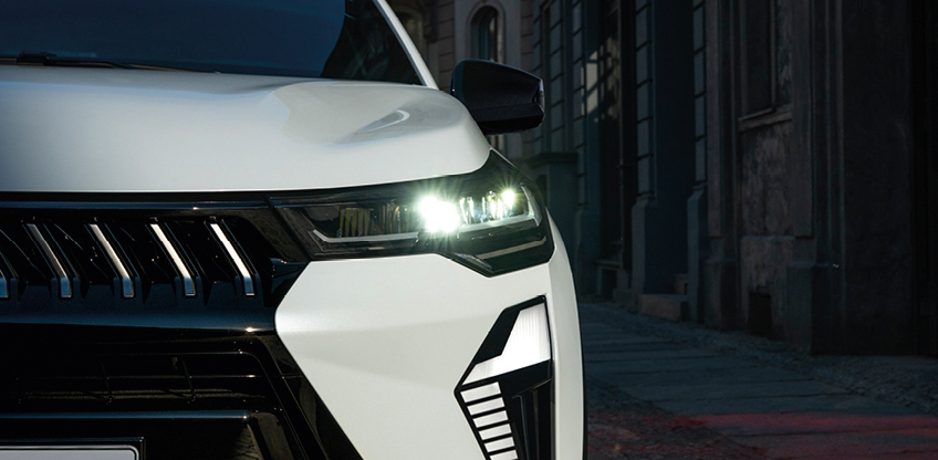 Detailaufnahme der Front eines wei&szlig;en Mitsubishi Grandis SUV in der Stadt, mit eingeschalteten LED-Scheinwerfern und markantem Tagfahrlicht-Design.