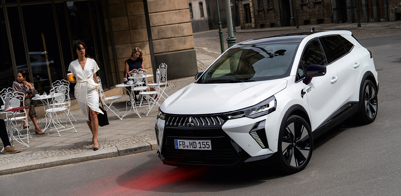 Wei&szlig;er Mitsubishi Grandis SUV in der Stadt vor einem Stra&szlig;encaf&eacute;, Frau in wei&szlig;em Kleid mit Kaffeebecher geht am Fahrzeug vorbei