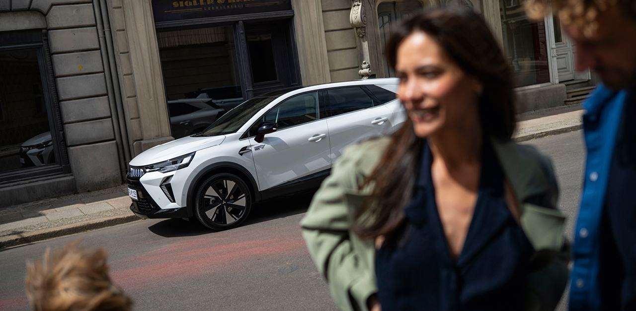 Wei&szlig;er Mitsubishi Grandis SUV vor einem historischen Stadtgeb&auml;ude, im unscharfen Vordergrund eine lachende Frau in Bewegung.