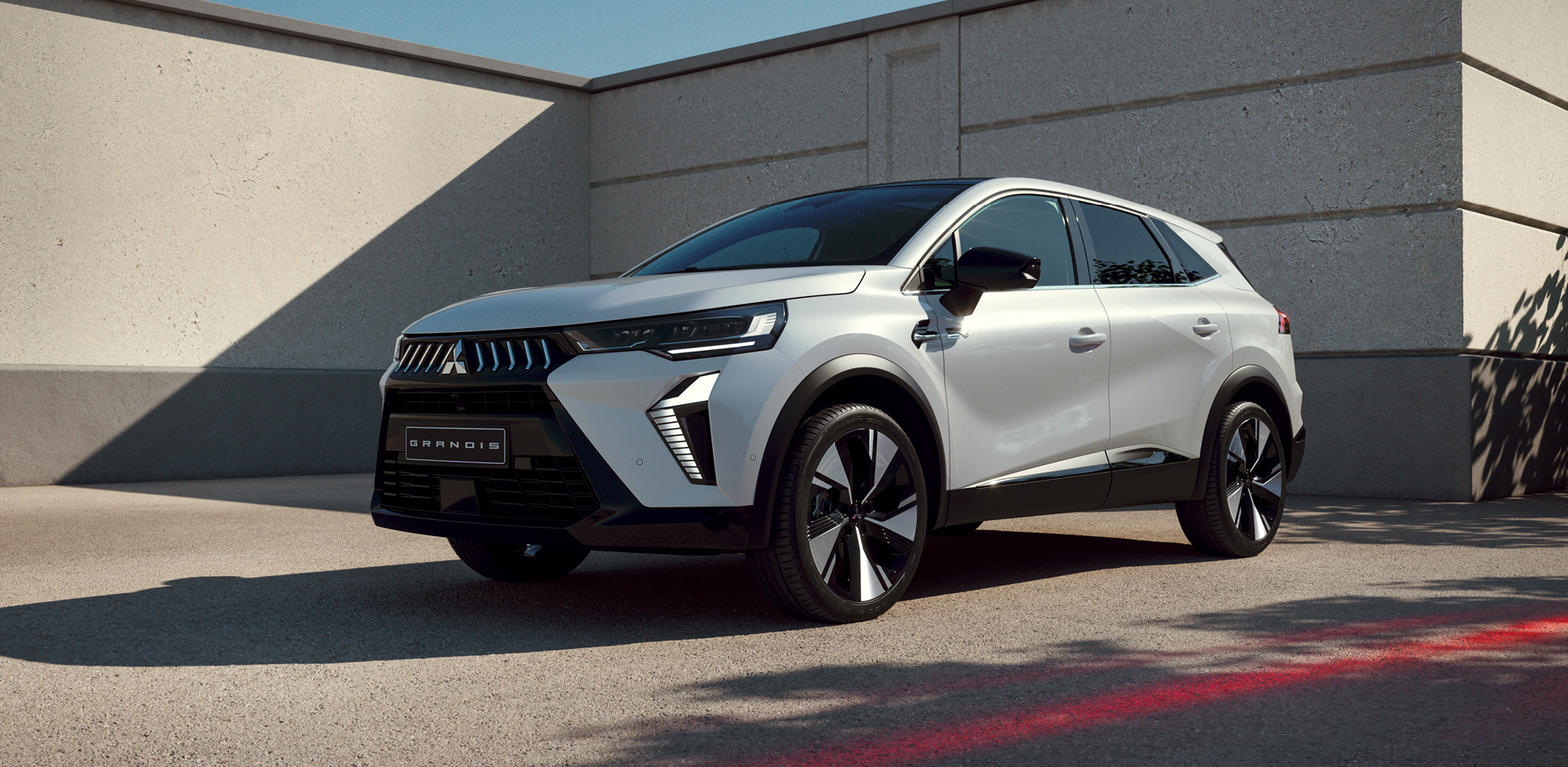 Wei&szlig;er Mitsubishi Grandis steht im Dreiviertel-Profil vor einer modernen Betonwand im Sonnenlicht, mit Fokus auf markanter Frontpartie und auff&auml;lligem Felgendesign.