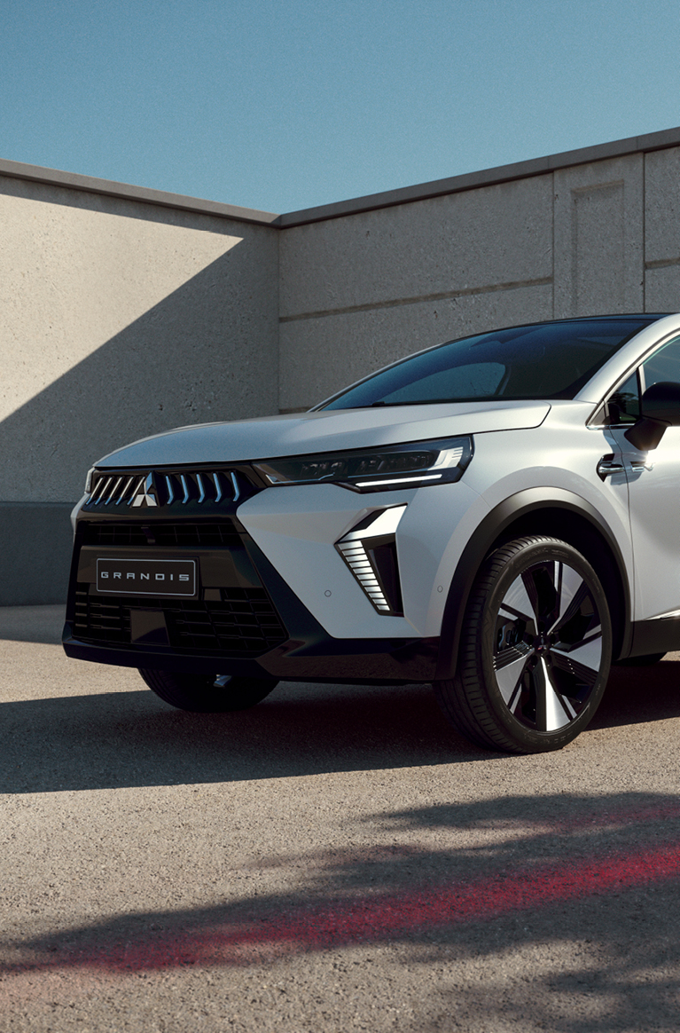 Wei&szlig;er Mitsubishi Grandis steht im Dreiviertel-Profil vor einer modernen Betonwand im Sonnenlicht, mit Fokus auf markanter Frontpartie und auff&auml;lligem Felgendesign.