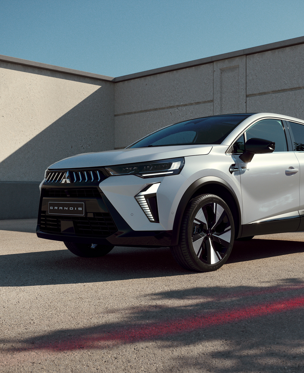 Wei&szlig;er Mitsubishi Grandis steht im Dreiviertel-Profil vor einer modernen Betonwand im Sonnenlicht, mit Fokus auf markanter Frontpartie und auff&auml;lligem Felgendesign.