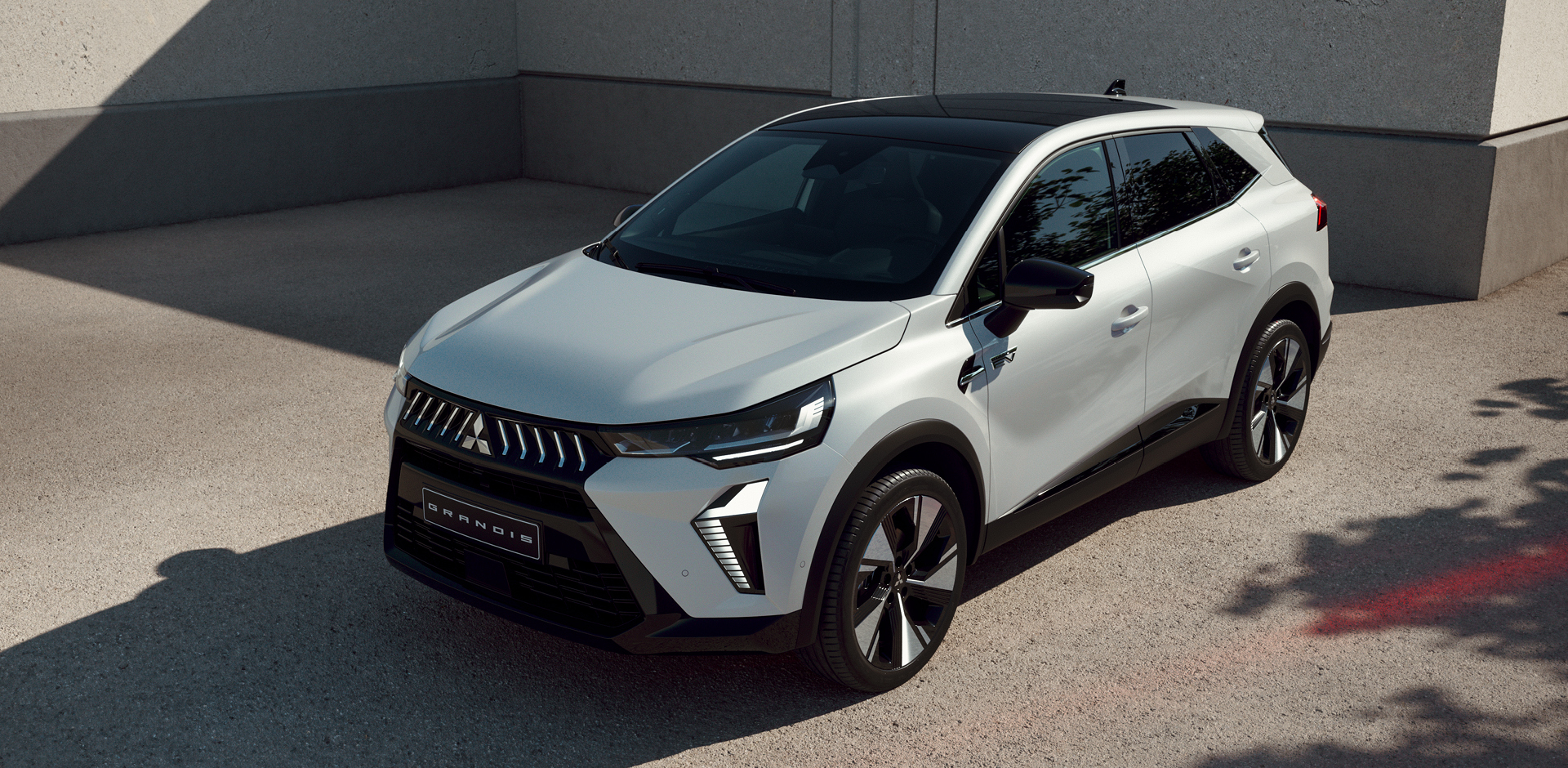 Wei&szlig;er Mitsubishi Grandis Kompakt-SUV steht vor einer Betonwand im Sonnenlicht, mit Fokus auf der modernen Frontpartie.