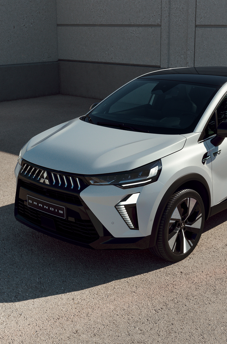 Wei&szlig;er Mitsubishi Grandis Kompakt-SUV steht vor einer Betonwand im Sonnenlicht, mit Fokus auf der modernen Frontpartie.