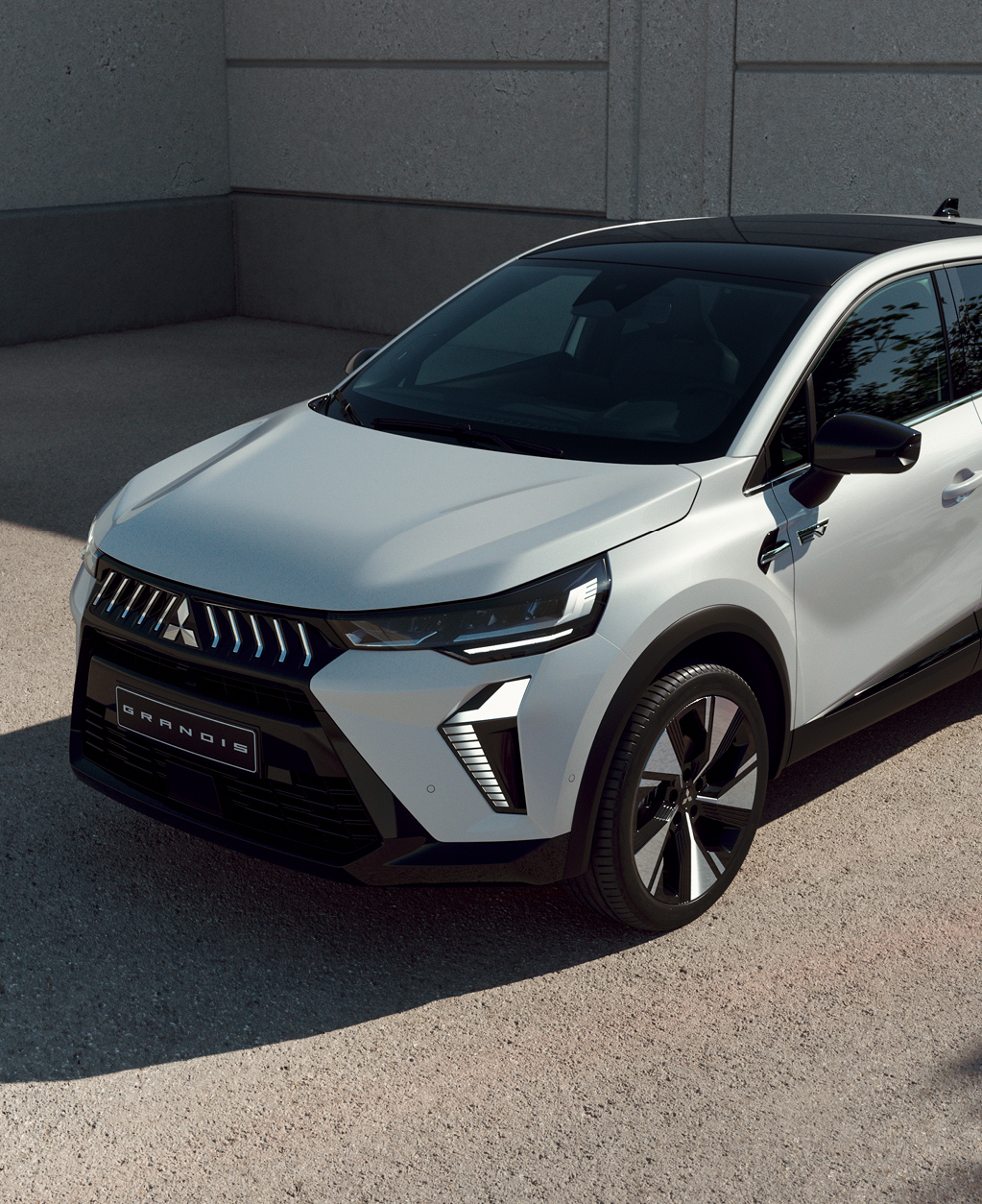 Wei&szlig;er Mitsubishi Grandis Kompakt-SUV steht vor einer Betonwand im Sonnenlicht, mit Fokus auf der modernen Frontpartie.